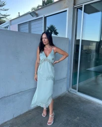Woman wearing a sage green silk slip dress with lace trim standing outdoors against a concrete wall and glass door.