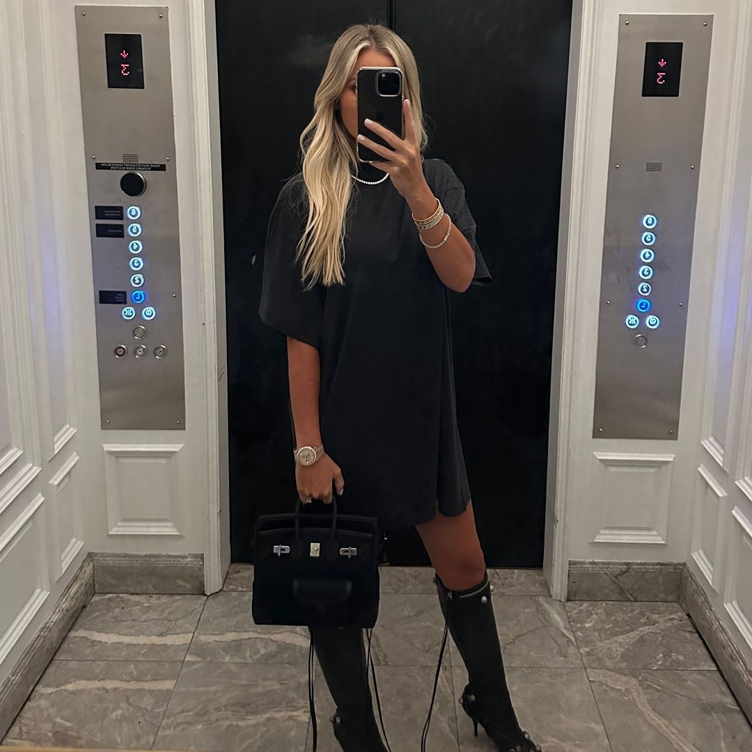 Blonde woman in black T-shirt dress and black heeled boots poses for mirror selfie in elevator holding black handbag.