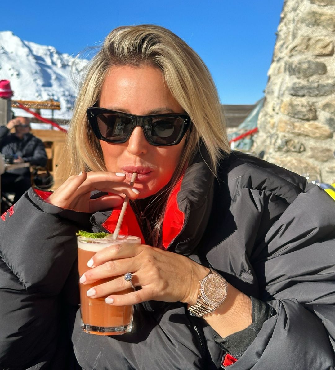 Woman in a black puffer jacket and sunglasses sipping a cocktail on a sunny day near snow-capped mountains at an outdoor cafe.