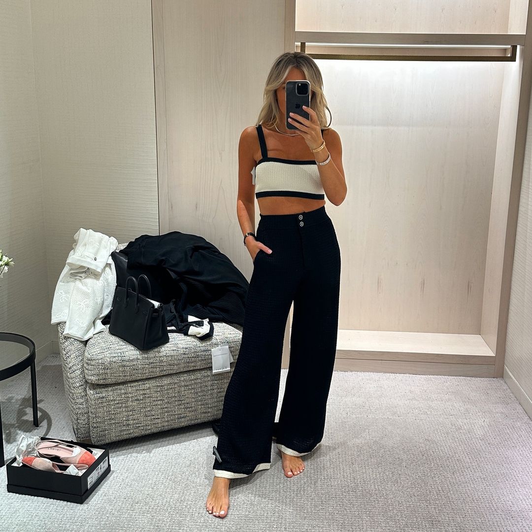 An adult female poses in a dressing room, wearing a crop top and wide-leg pants while holding a smartphone for a mirror selfie.
