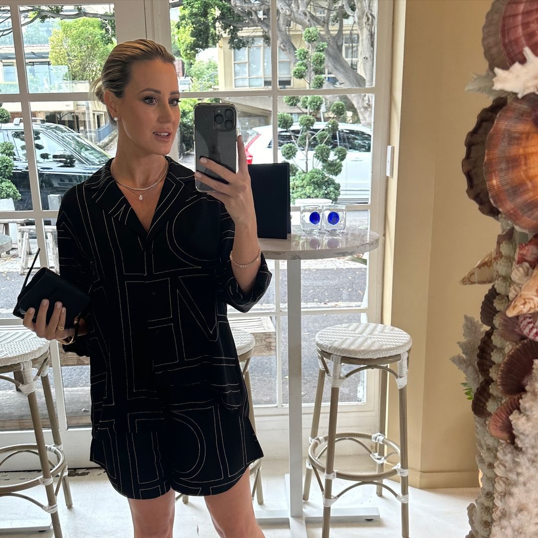 Blond woman in black printed shorts suit takes a selfie indoors, holding a black clutch bag near a decorative seashell pillar.