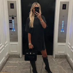 Blonde woman taking mirror selfie in elevator wearing black oversized t-shirt, tall boots, and holding black handbag.