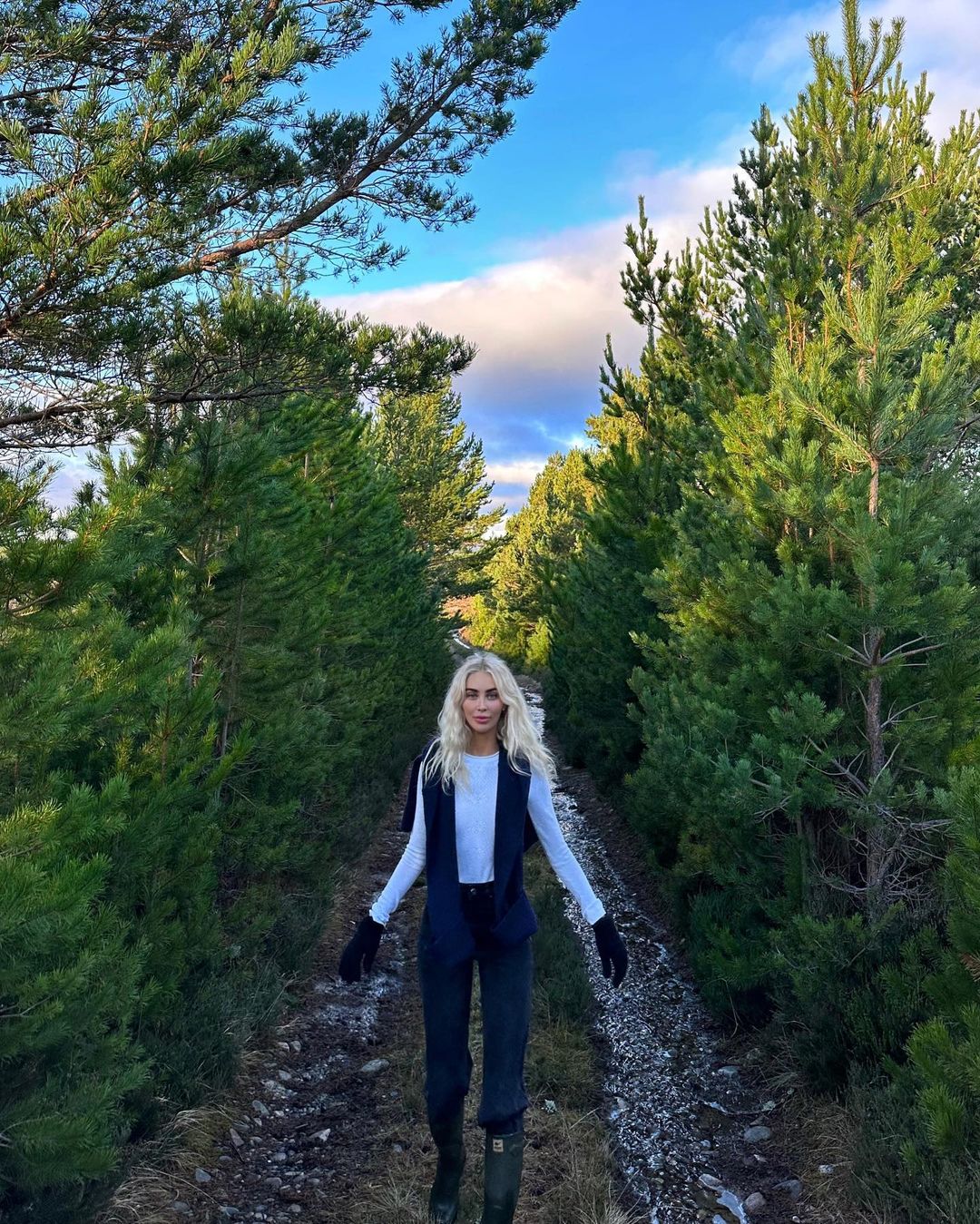 A woman in a white top and dark vest walks on a dirt path flanked by tall green pine trees under a blue sky with wispy clouds.