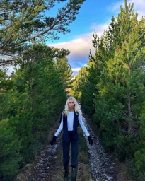 Blonde woman walking along a dirt path lined with pine trees wearing a white top, dark trousers, and dark vest.
