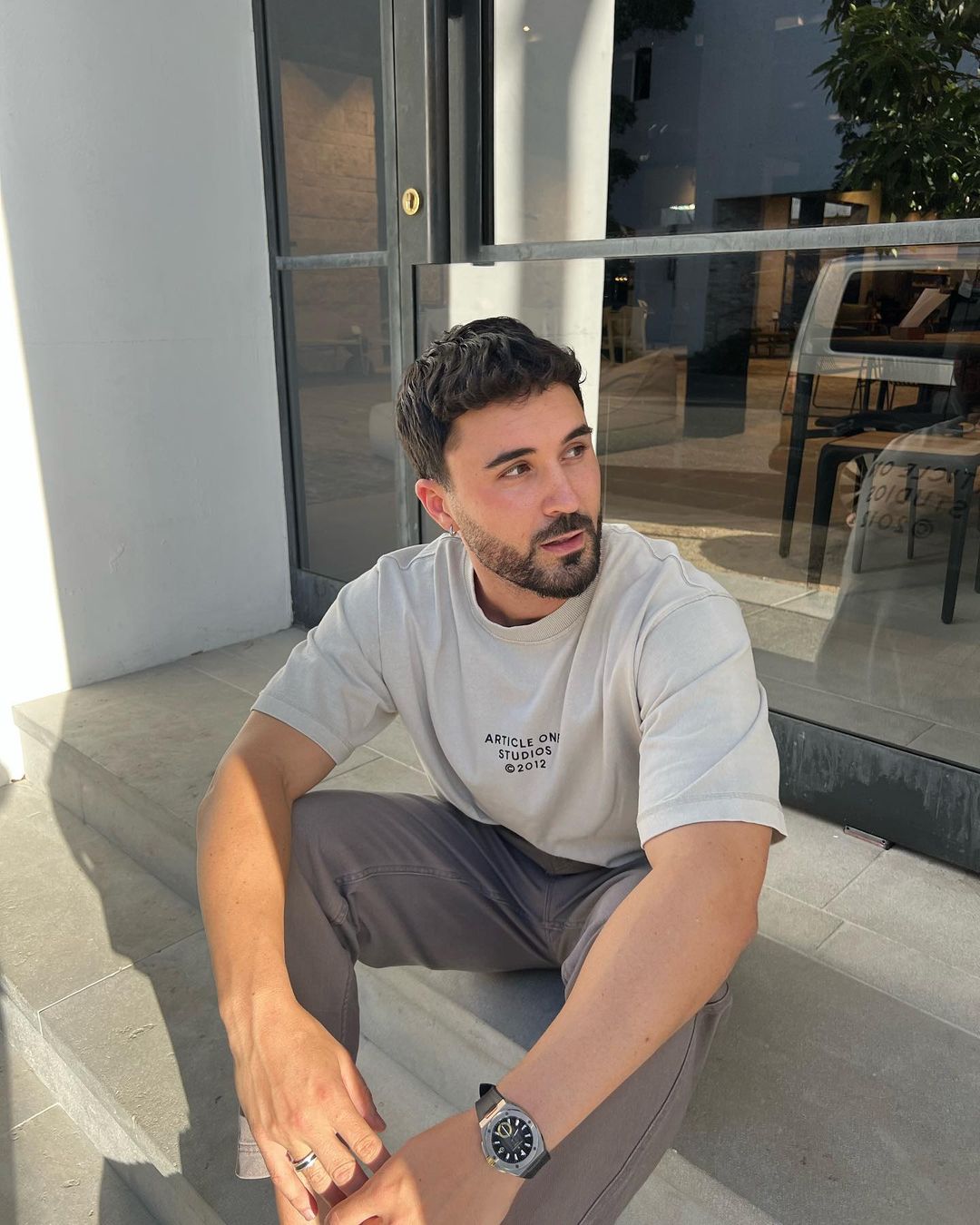 A young adult male with a beard is sitting on concrete steps outside a building with glass doors, wearing a t-shirt, pants, and a wristwatch.