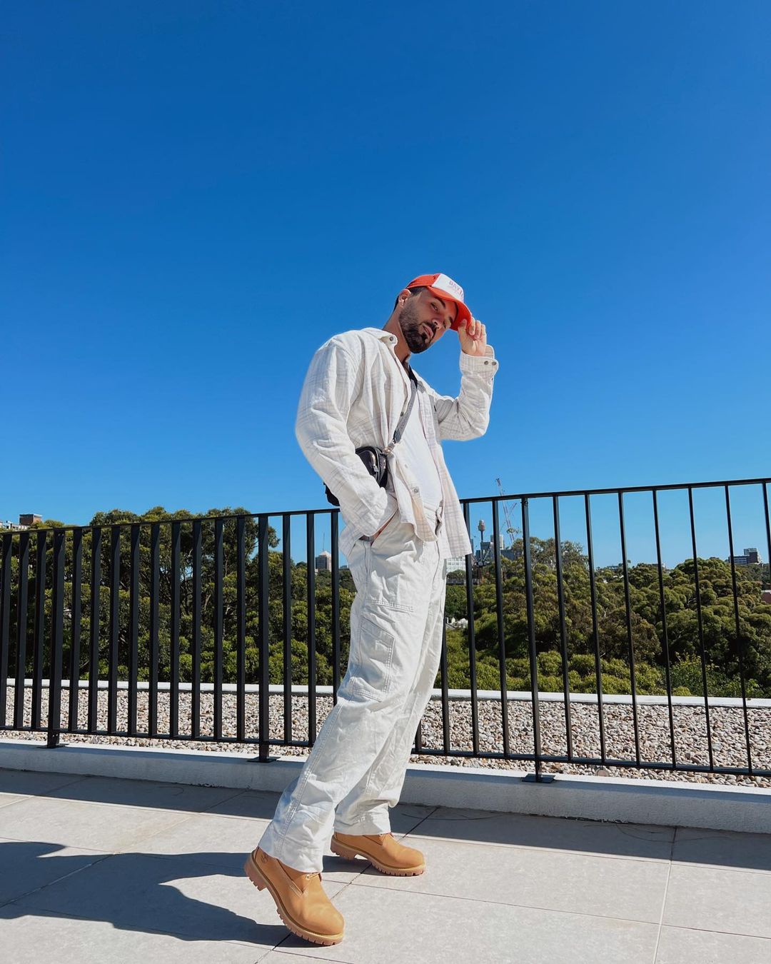 Bearded adult male posing in outdoor setting wearing matching off-white jacket and pants, tan ankle boots, and orange and white baseball cap.
