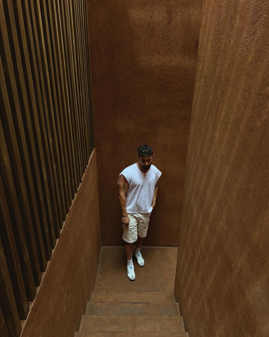 Man walking down staircase with brown walls and wood slat accent. Wearing a white tank top, shorts, and sneakers in interior setting.