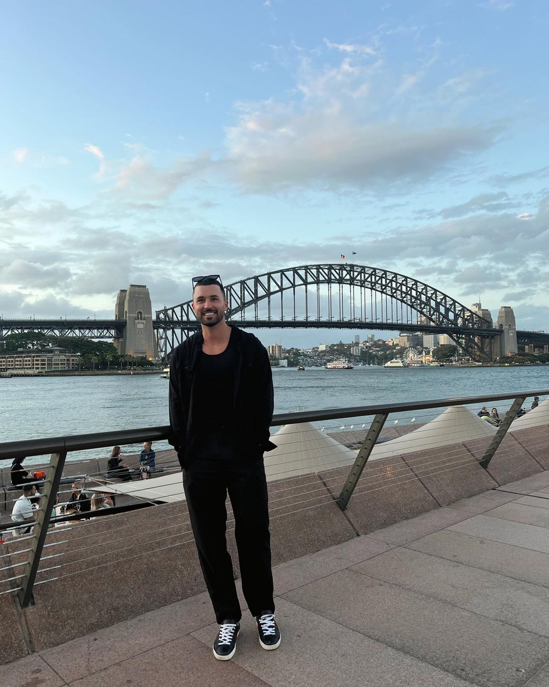Adult male in black casual clothes posing near Sydney Harbour Bridge with cloudy sky and waterfront views in the background on a sunny day.