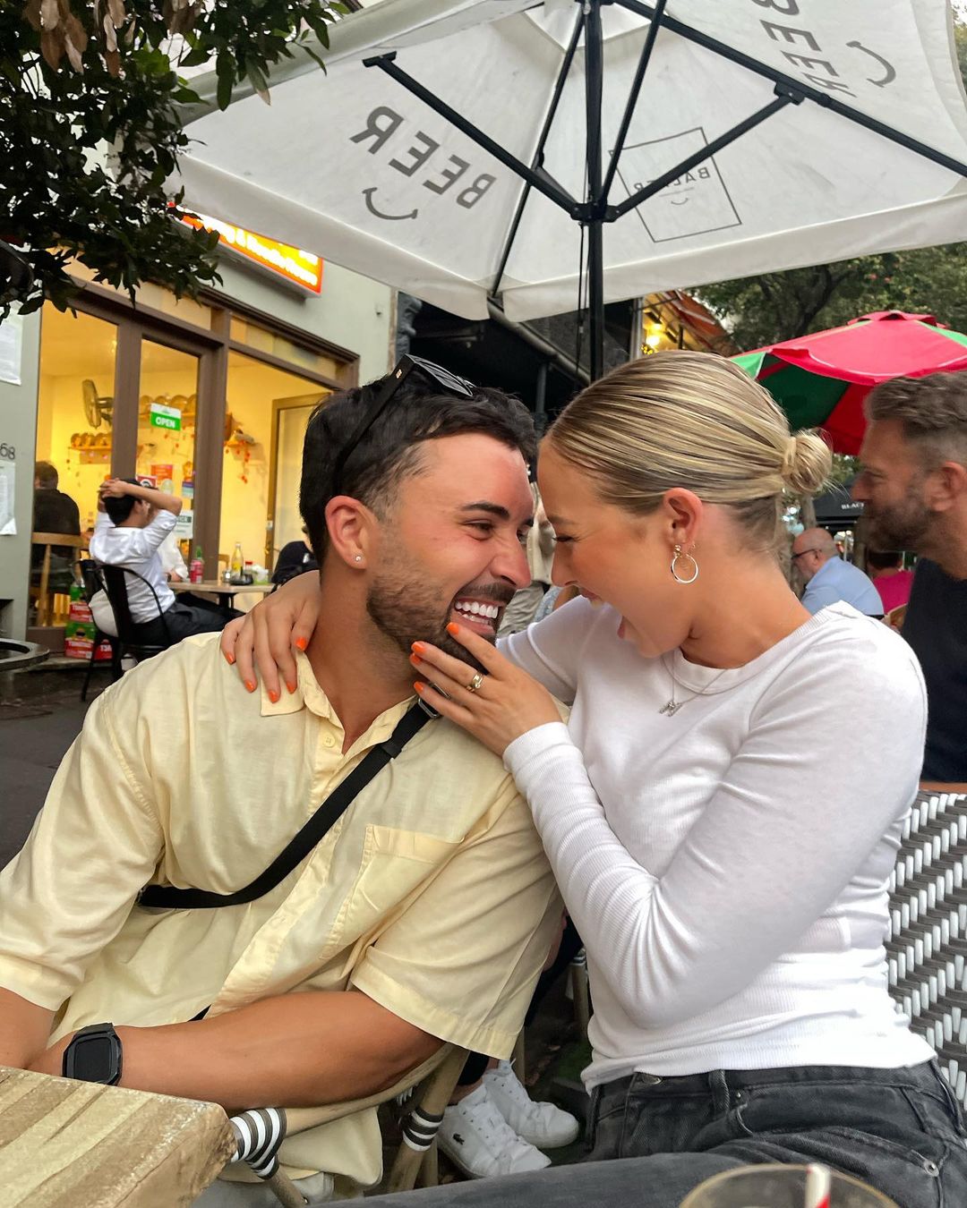 A smiling couple embraces at an outdoor cafe with white umbrella and tables, captured in a medium shot during daylight.