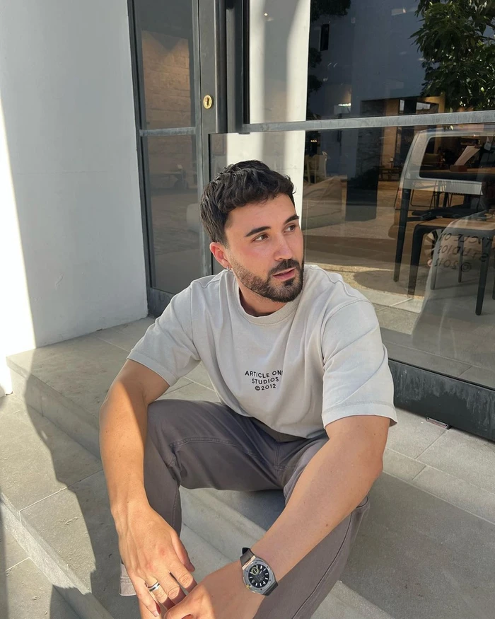 Man with beard wearing an off-white t-shirt and grey trousers sitting on stone steps outdoors.