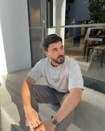Man with beard wearing an off-white t-shirt and grey trousers sitting on stone steps outdoors.