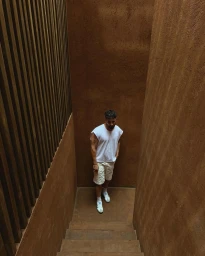 Man in white t-shirt and shorts walking down textured brown stairs between vertical wooden slats.