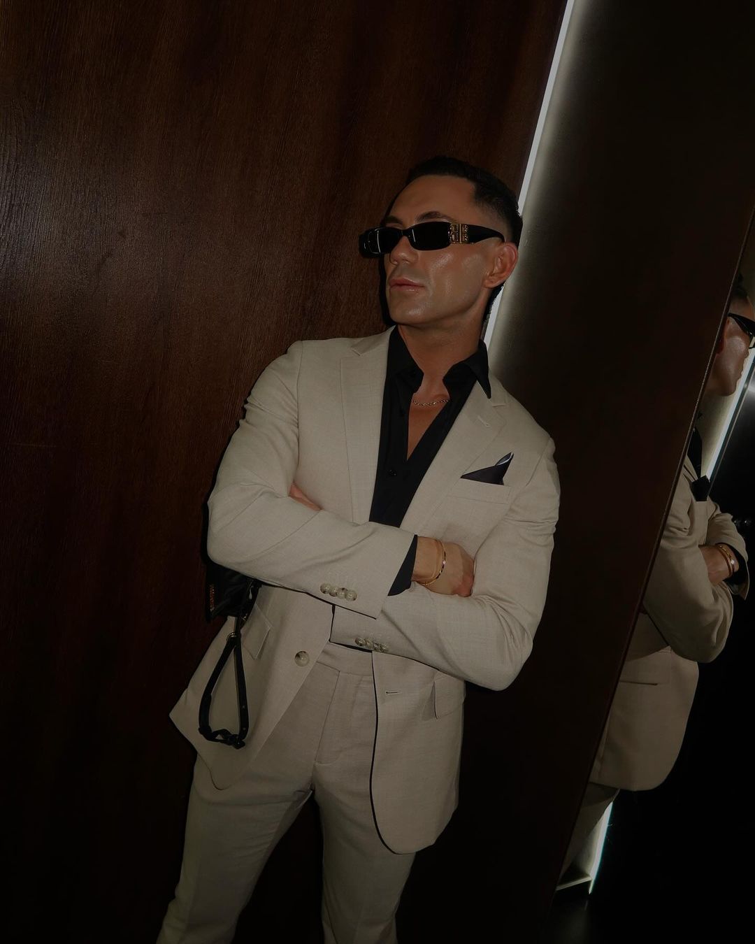A man in a beige suit and sunglasses stands indoors with arms crossed, leaning against a wall, with a neutral expression on his face.