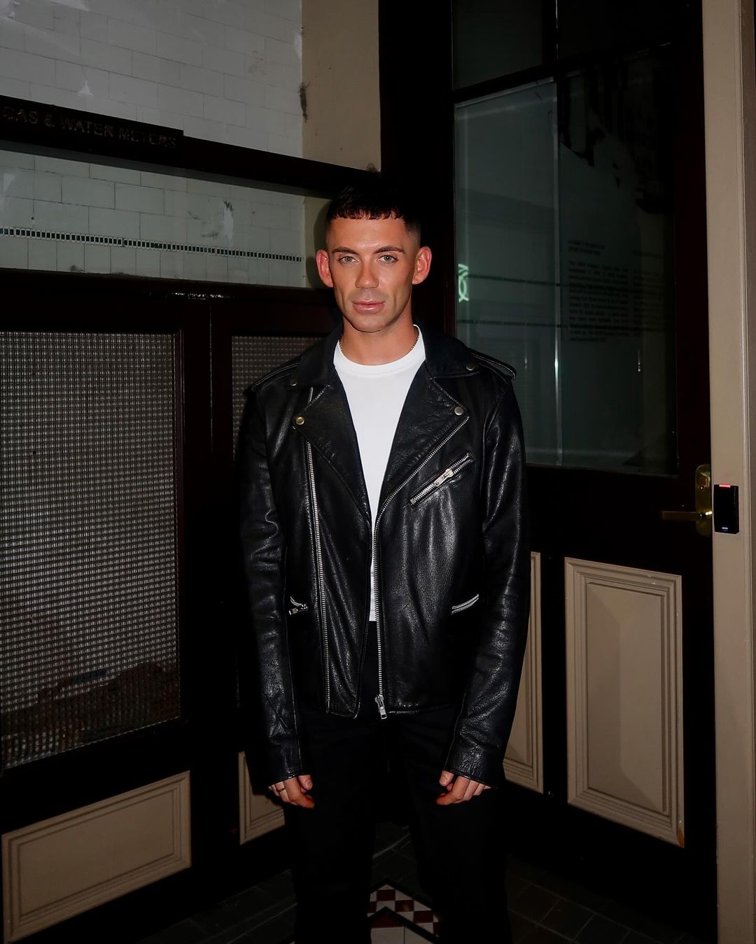 Stylish man in a black leather jacket and white t-shirt poses in front of a dark door. Fashion-forward and confident pose.