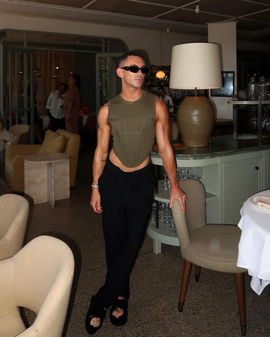 Man poses in olive tank top and black pants in restaurant. Stylish sunglasses, bracelets, and neutral decor complete the scene.