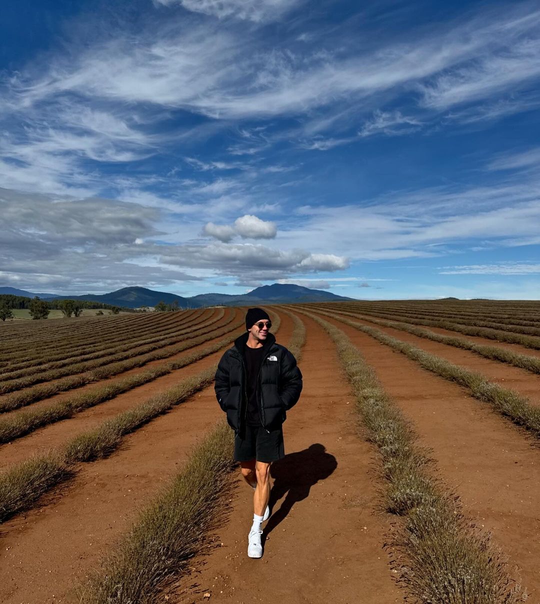 An adult male strolls in a black puffer jacket and shorts on a dirt path lined with lavender bushes beneath a blue sky.