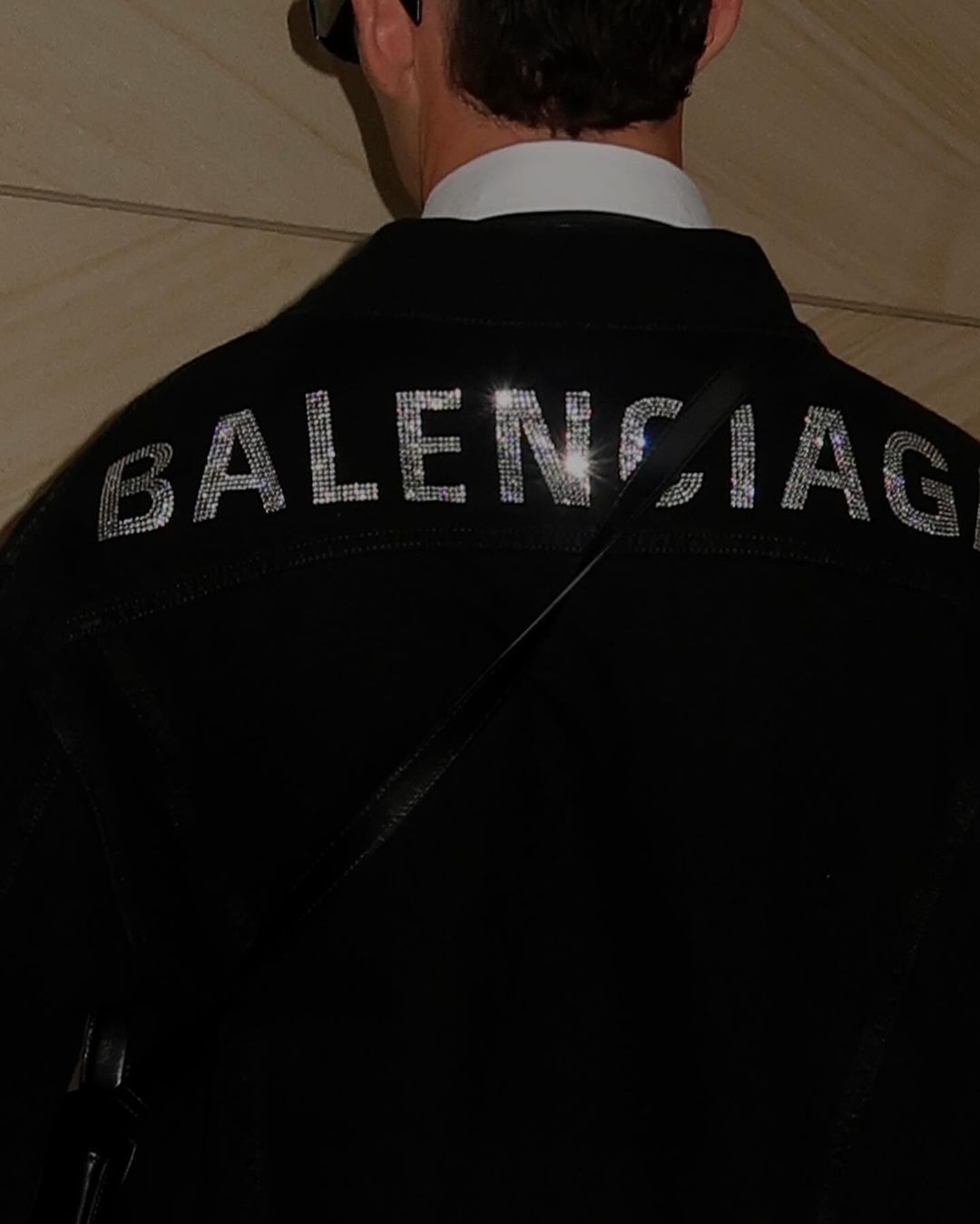 Rear view of a person in a black jacket with 'Balenciaga' spelled out in rhinestones, accessorized with a black bag and sunglasses.