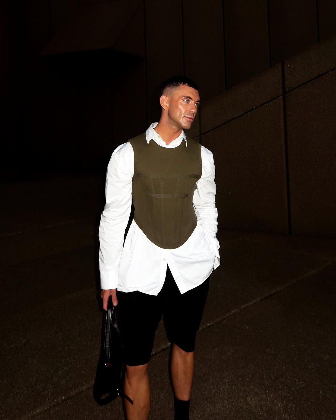 Man in modern olive vest and black shorts poses outdoors against a textured wall, holding a black purse, capturing a unique fashion statement.