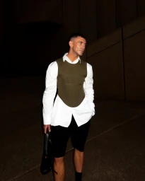 Man wearing an olive green corset vest over a white button-down shirt and black shorts holding a black bag