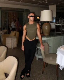 Man wearing sunglasses, olive green sleeveless top, and black trousers posing in a restaurant interior by Jordan Kay