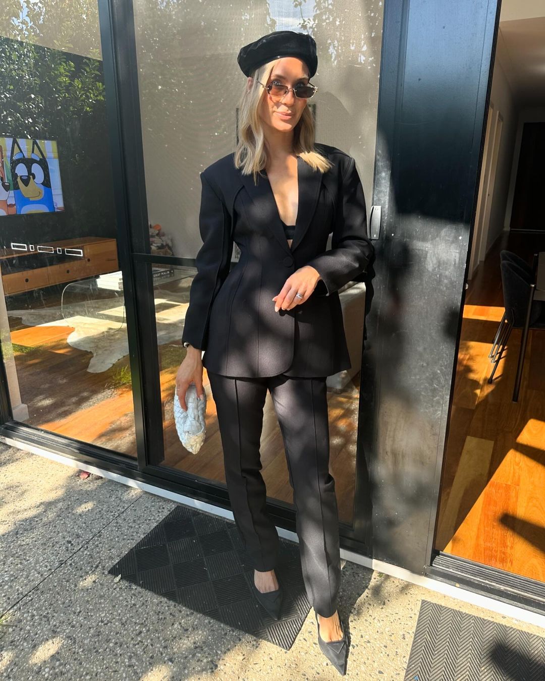 Woman in black suit and beret stands outdoors near a glass wall, holding a beaded clutch purse, fashionable and chic in sunlight.