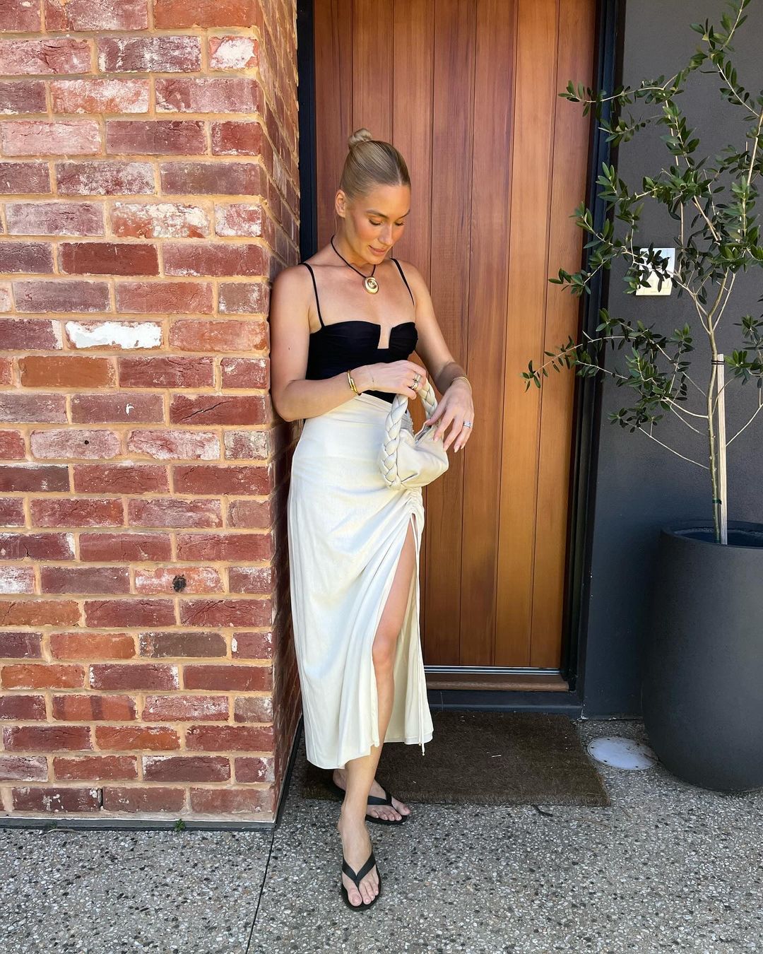 A woman in a black top and ivory skirt poses against a brick wall next to a dark door, holding a beige woven handbag.