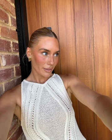 Woman in a white sleeveless knitted top posing outdoors against a brick wall and wooden door, photographed by Jamie Reilly.