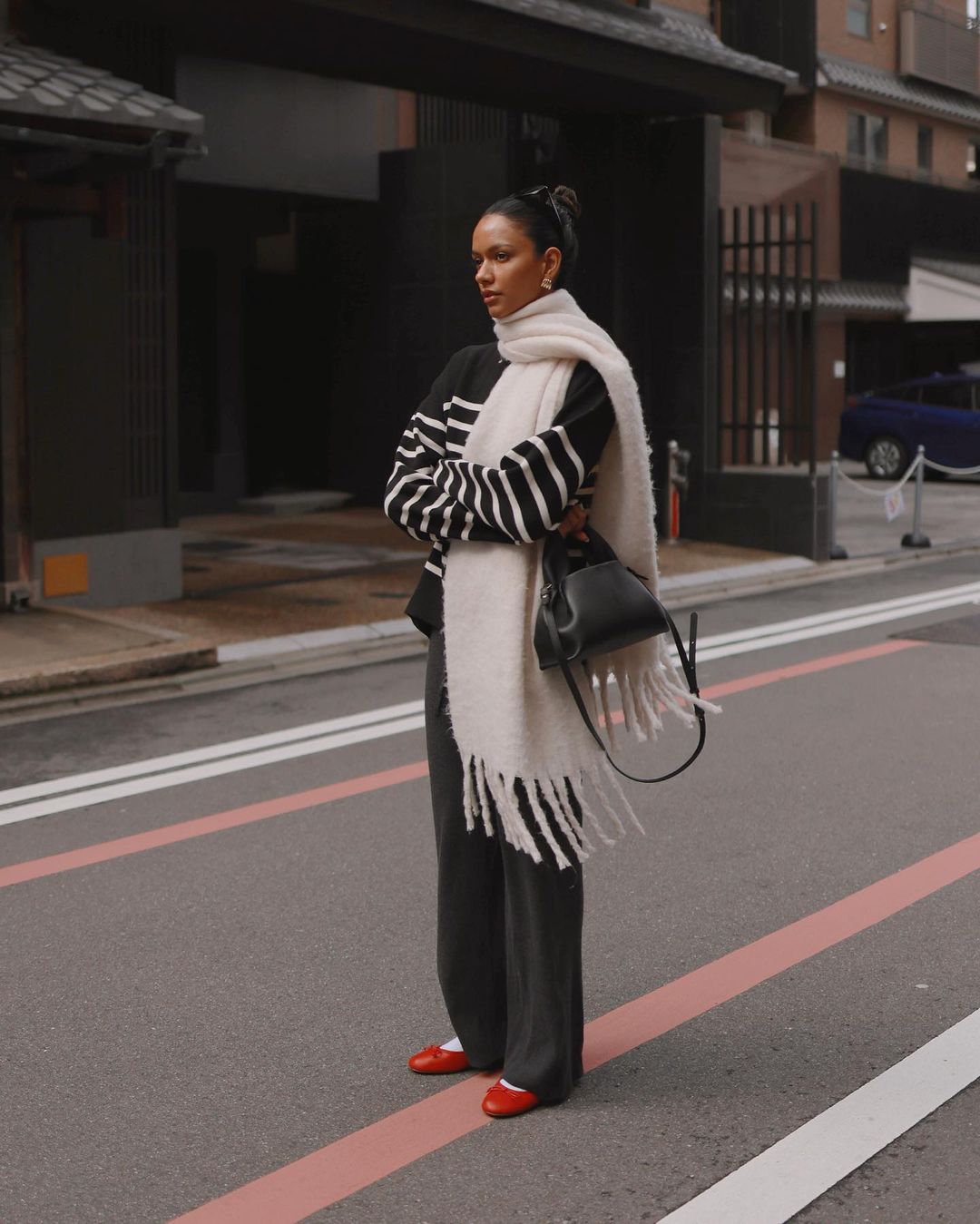 Adult female in black and white striped sweater, oversized scarf, and red shoes standing on a city street with buildings in the background.