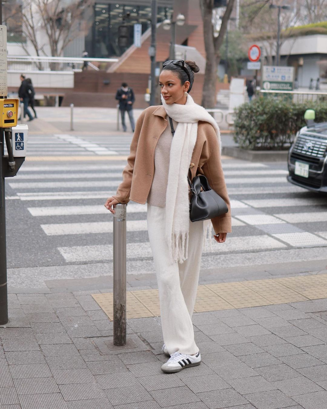A woman in a tan jacket and cream-colored pants stands on a city crosswalk, wearing Adidas sneakers and a fringed scarf.
