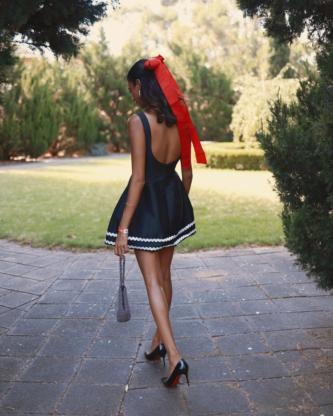 Woman in a black dress with a red ribbon walks on a stone path in a green outdoor setting, carrying a silver handbag and wearing black high heels.