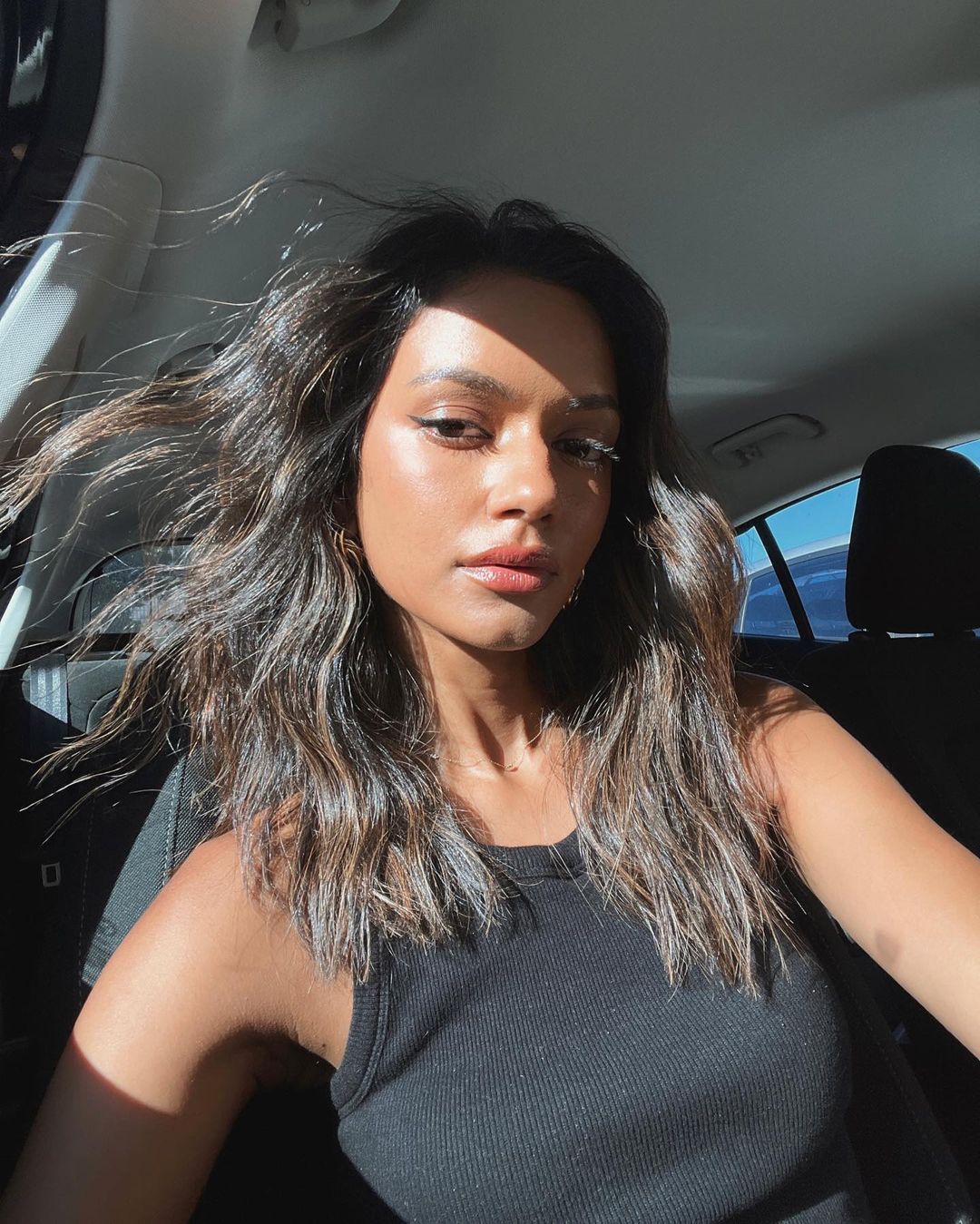 Portrait of a tanned woman with wavy brunette hair, wearing a black tank top, inside of a car with sunlight coming through window.