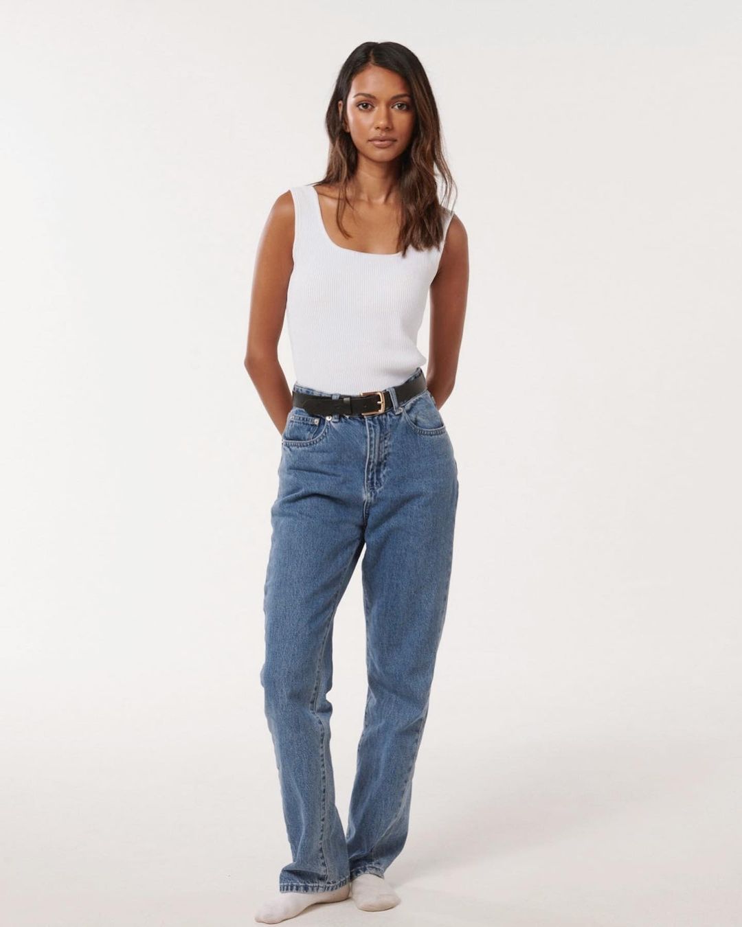 A woman in a white tank top and blue jeans stands against a white backdrop in a fashion-style, full-length studio shot.