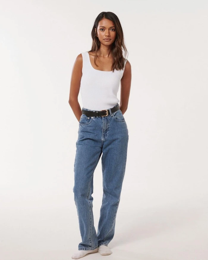 A woman wearing a white tank top and blue jeans stands against a plain light background.