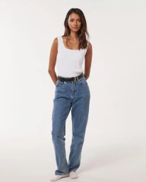 A woman wearing a white tank top and blue jeans stands against a plain light background.