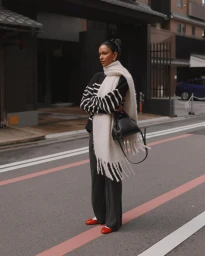 Woman in a striped sweater and long cream scarf holding a black handbag standing on an urban street.