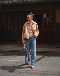 Woman walking on a street wearing a tan jacket, striped turtleneck, blue jeans, and holding a black handbag.