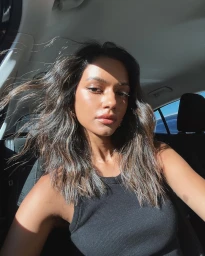 A woman with wavy brown hair wearing a black tank top taking a selfie inside a car.