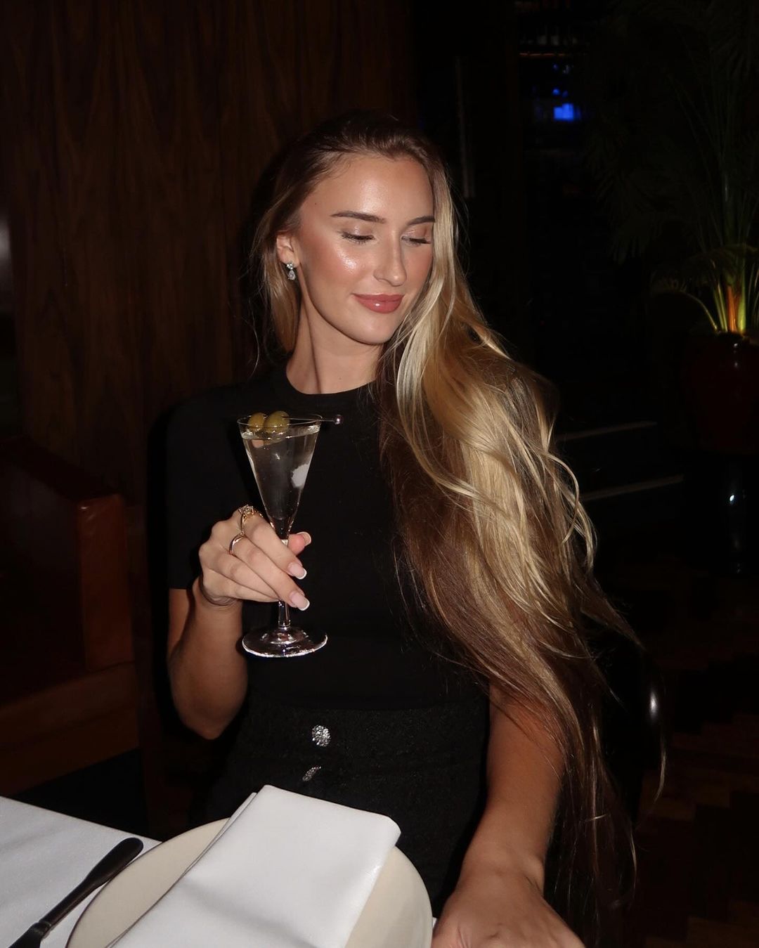 Adult woman in a dark restaurant is holding a martini glass with olives. She has long blond hair and is wearing a black dress.