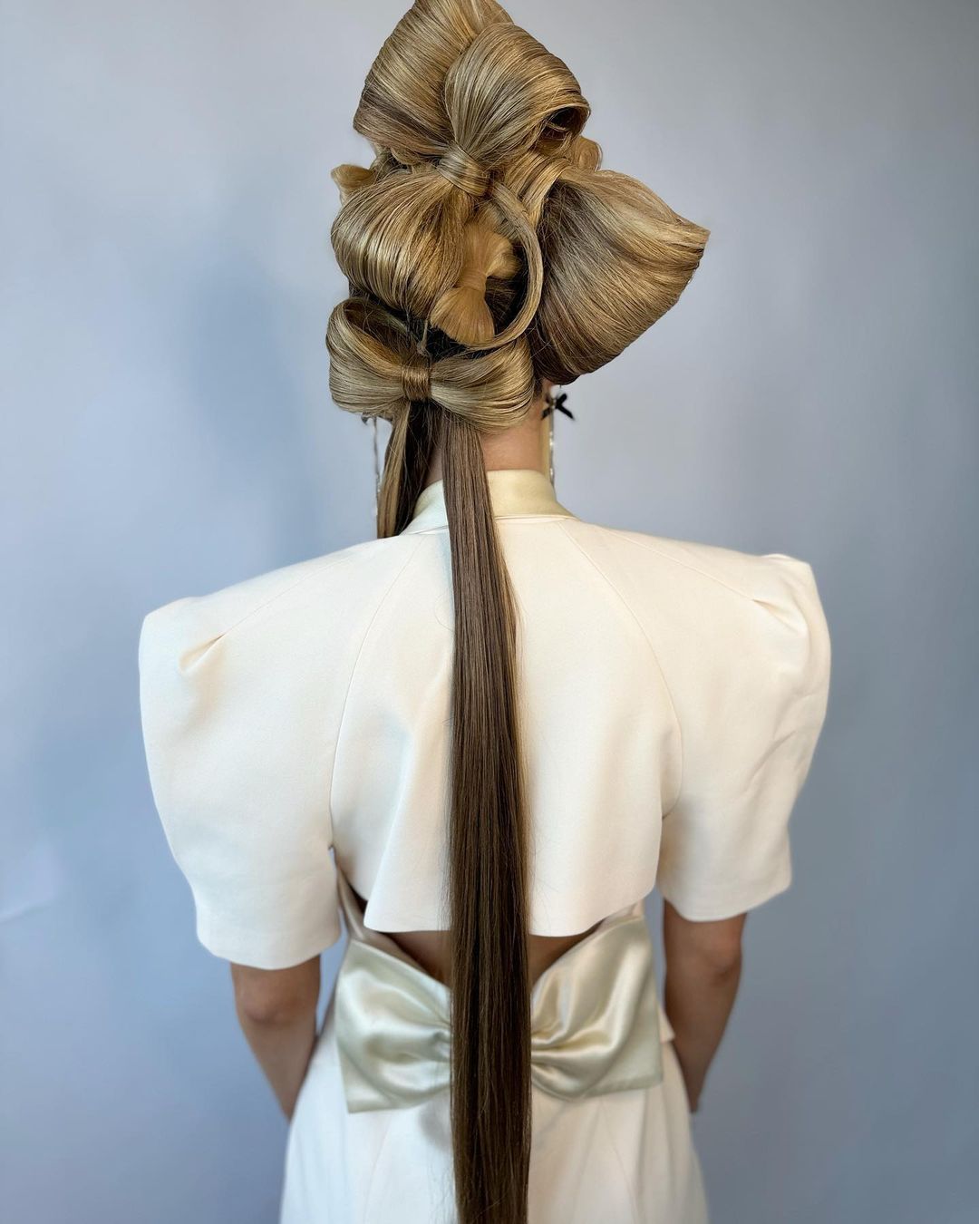 Blonde hair styled into a series of decorative bows, viewed from behind, with a woman wearing a cream-colored dress with back bow detail.