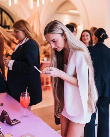 Young woman with long blonde hair applies makeup at a table, holding a compact mirror with beauty products and a cocktail glass nearby.