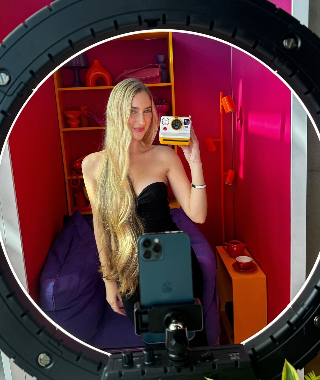 Blonde woman in a black dress poses with a yellow instant camera, framed by a ring light, in front of a colorful background.