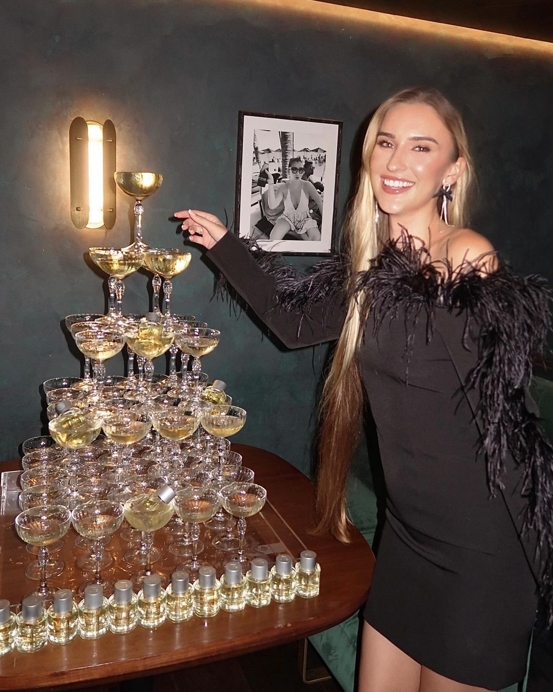 Smiling woman in a black dress with feather trim stands next to a champagne tower and liquor bottles on a wood table in a bar.
