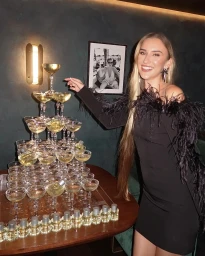 Smiling woman in a black feather-trimmed dress stands beside a large champagne glass tower on a dark wooden table.