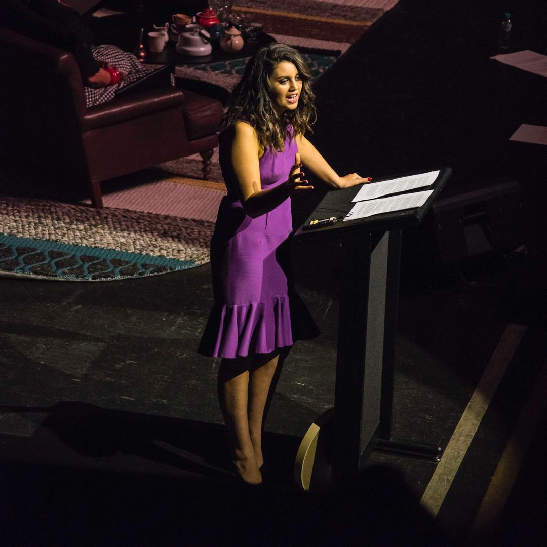 Woman in purple dress speaks at podium with papers in dark event space with rug, chair, and table visible in the background.