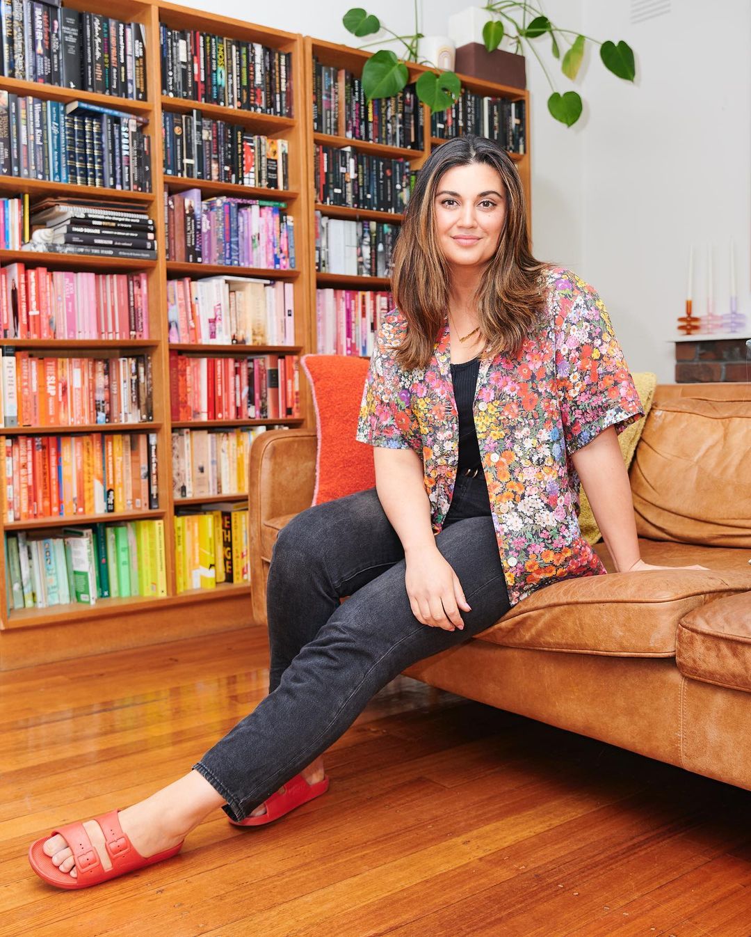 Woman sitting on a leather couch in a home interior with a bookshelf, wearing a floral short-sleeved shirt, jeans, and sandals.