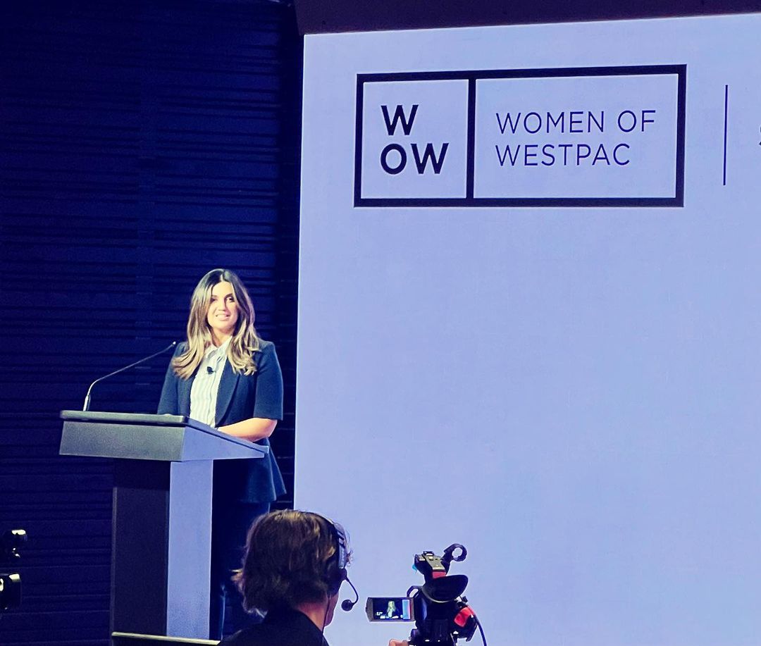Woman in business attire presents at a podium with 'Women of Westpac' logo, captured at a medium shot with a blue background.