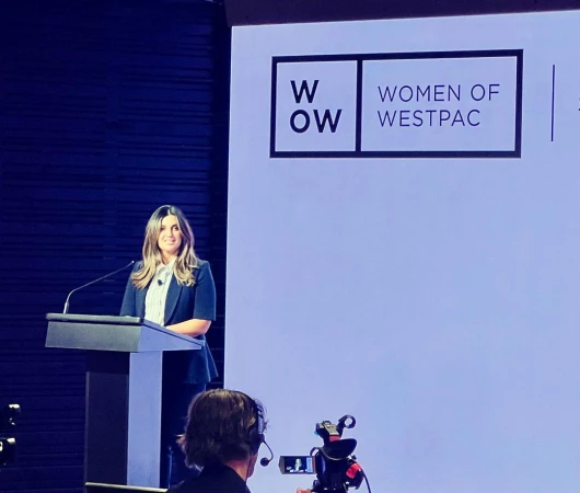 Woman in business attire presents at a podium with 'Women of Westpac' logo, captured at a medium shot with a blue background.
