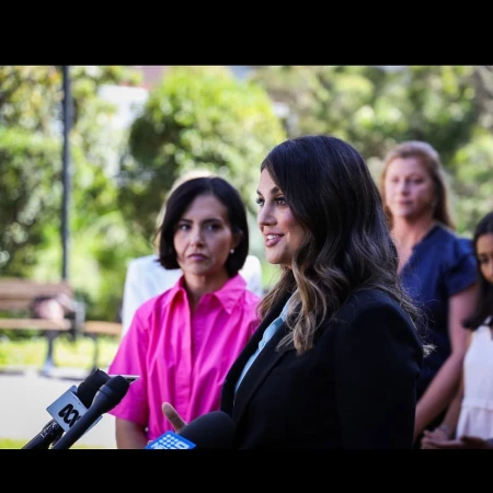 Brunette woman in a suit speaks at an outdoor press conference with microphones and other women in the background on a sunny day.