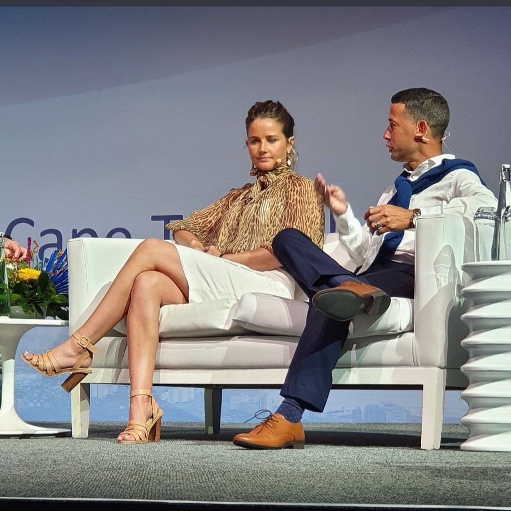 Two people, an adult woman and an adult man, sit on a white couch during a panel discussion at an indoor event.