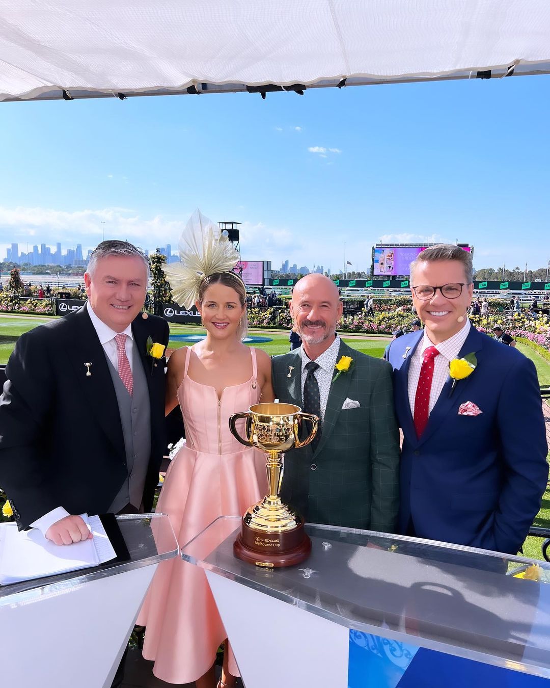 Four people pose behind a trophy at an outdoor event on a sunny day with a city skyline in the background. The trophy is gold and brown.